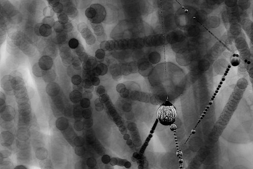 Ein unscharfes, monochromes Bild mit vielen kreisförmigen Formen, die in verschiedenen Größen und Hell-Dunkel-Kontrasten angeordnet sind. Im Vordergrund hängt eine auffällige, runde Perle oder Kugel, die durch ihre Textur hervorsticht. Die Umgebung wirkt abstrahiert und verschwommen.